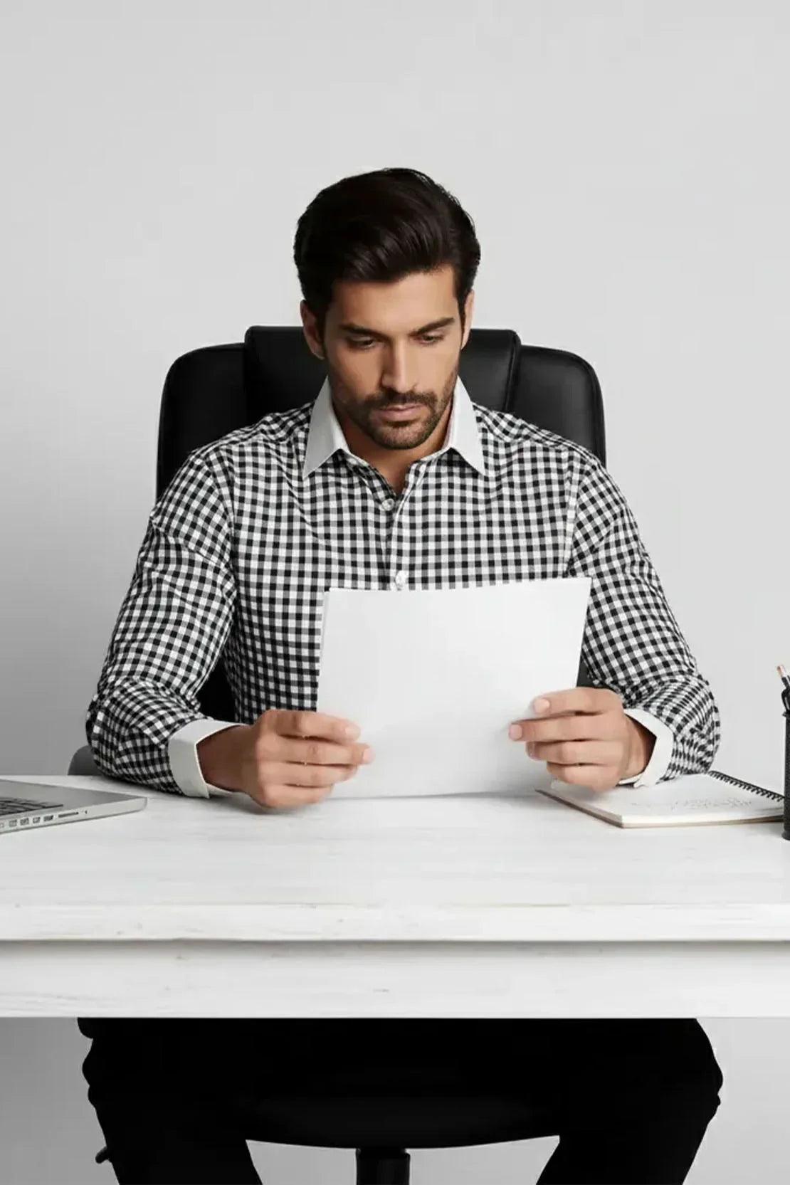 Man in black and white checkered shirt reading document at white office desk with laptop and notebook