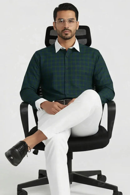 Man in green checked shirt with white collar and cuffs, white pants, black shoes, sitting on black office chair