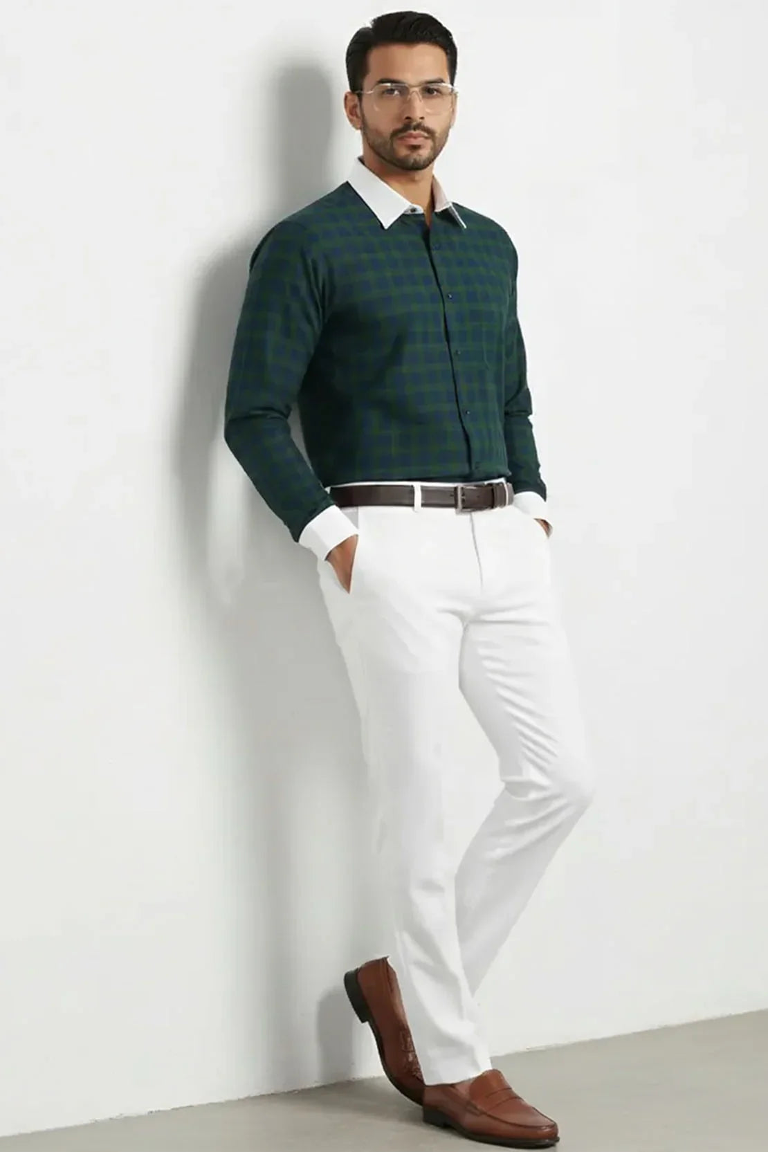 Man wearing green checkered formal shirt with white collar and cuffs, white trousers, brown belt, and brown shoes, standing against white wall