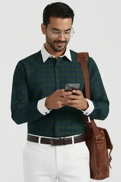 Man wearing green checkered shirt with white collar and cuffs, white pants, brown belt and brown leather shoulder bag, using smartphone
